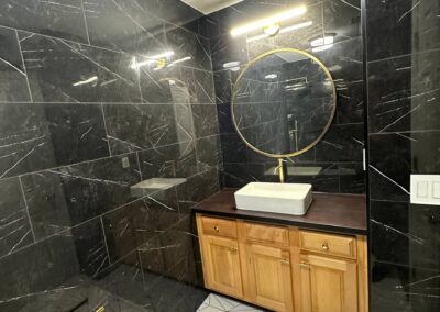 A remodeled bathroom with black marble tile walls featuring white veining, a modern bathtub, and a geometric-patterned floor. Gold fixtures, including a rainfall showerhead and hardware, enhance the luxurious and contemporary style.