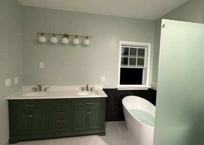 A remodeled bathroom with a green double-sink vanity, elegant lighting, a freestanding tub, and a frosted glass shower enclosure with pebble tile flooring, blending functionality and sophistication in a serene, contemporary design.