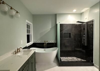 A beautifully remodeled bathroom with a sleek freestanding tub, a black-tiled walk-in shower with pebble flooring, and a green double-sink vanity. Modern lighting fixtures, updated hardware, and fresh flooring complete the sophisticated, contemporary design.