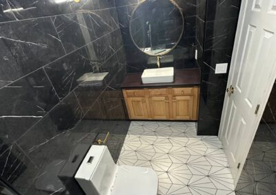 A luxurious bathroom featuring black marble walls with white veining, a geometric-patterned floor, a sleek vessel sink with gold fixtures, and a round mirror. A modern toilet and elegant lighting complete the sophisticated design.