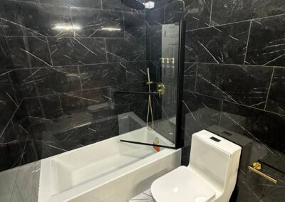 A modern bathroom with black marble tile walls featuring white veining, a sleek bathtub, and a geometric-patterned floor. Gold fixtures, including a rainfall showerhead and hardware, complement the luxurious and contemporary design.