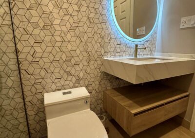 Powder room with a geometric tile accent wall, floating wood vanity, backlit LED circular mirror, and light wood flooring