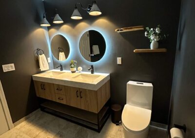 Modern bathroom with a double vanity, LED circular mirrors, dark accent wall, floating shelves, and light tile flooring.