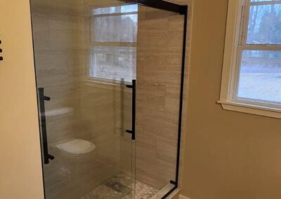 Walk-in shower with black-framed glass doors, wood-look tile walls, and pebble floor