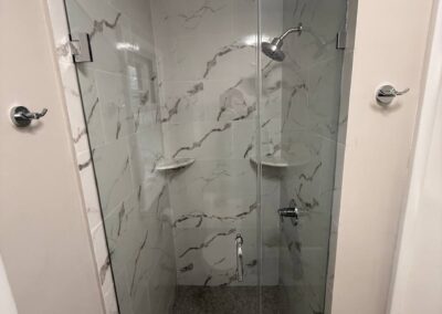 Walk-in shower with marble-look tiles, frameless glass door, and built-in corner shelves
